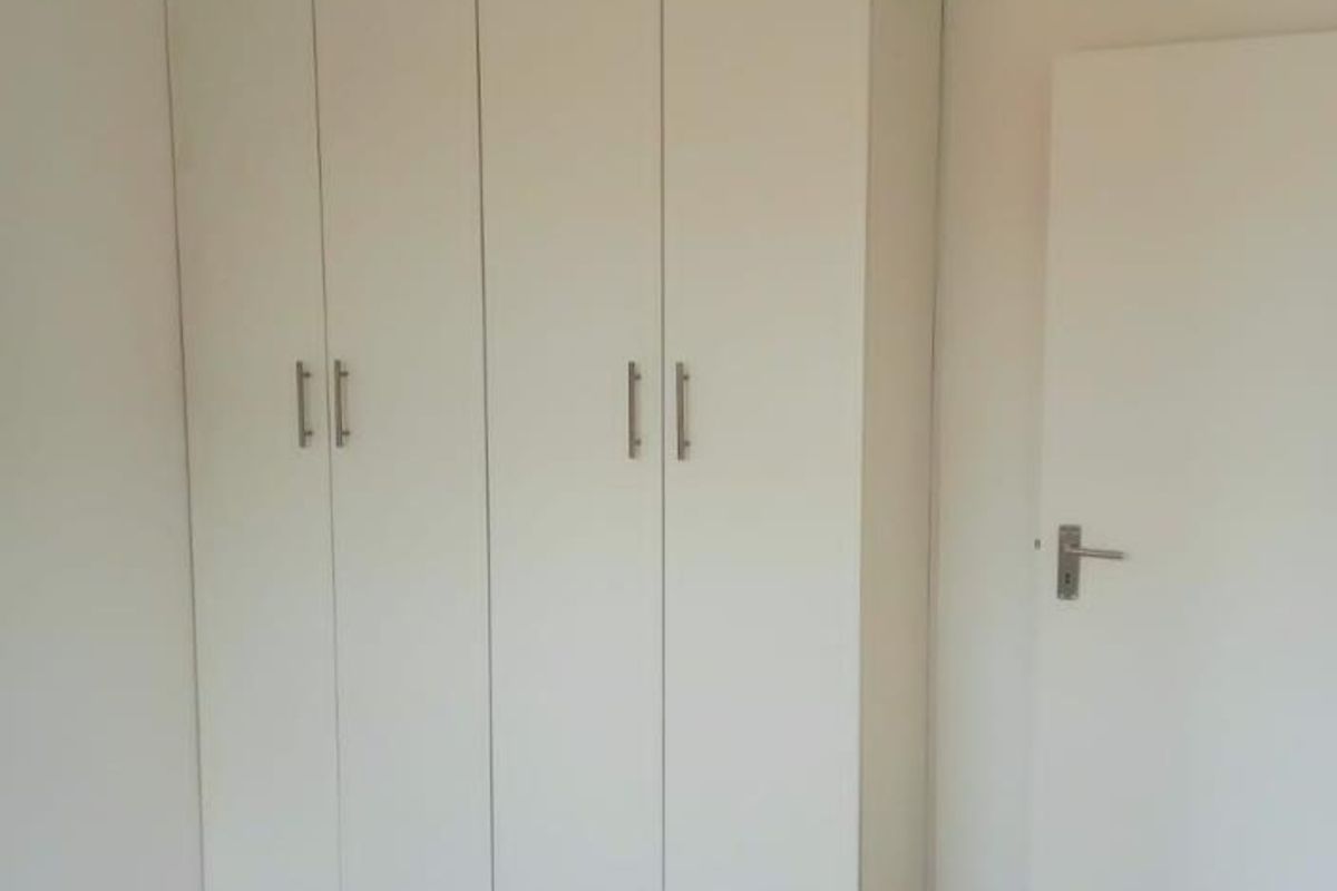 A clean, empty bedroom featuring neutral laminate flooring and a large, white built-in wardrobe with modern handles.