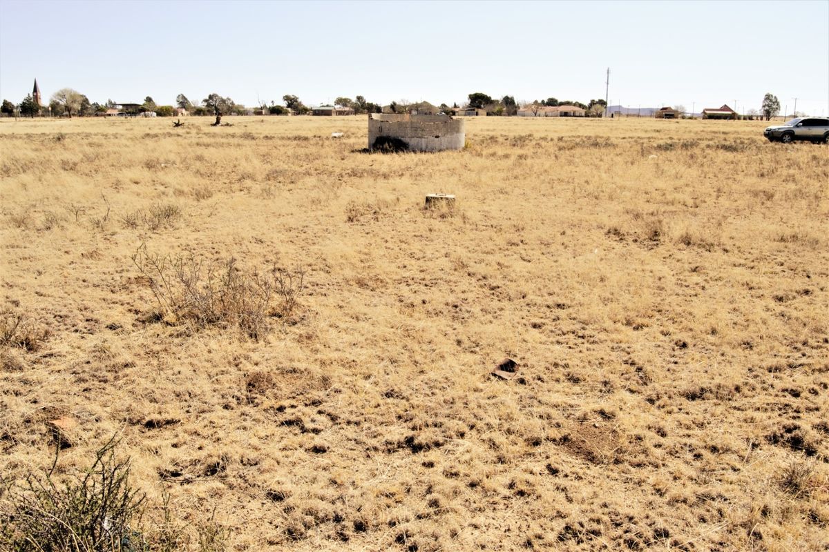 A wide view of a vacant residential plot featuring dry grass and a small, weathered concrete water reservoir structure.