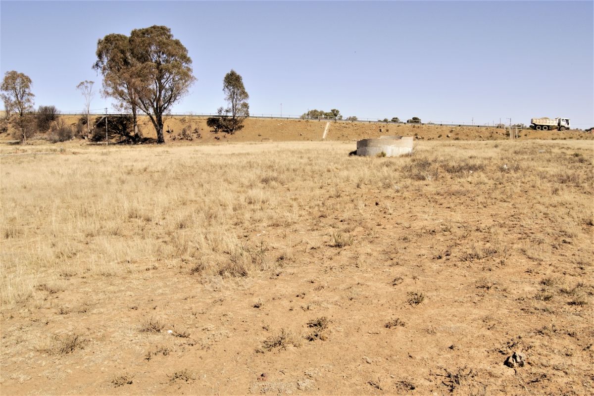 A wide-angle view of a vacant residential plot featuring dry grass, mature trees, and a small concrete water tank structure.