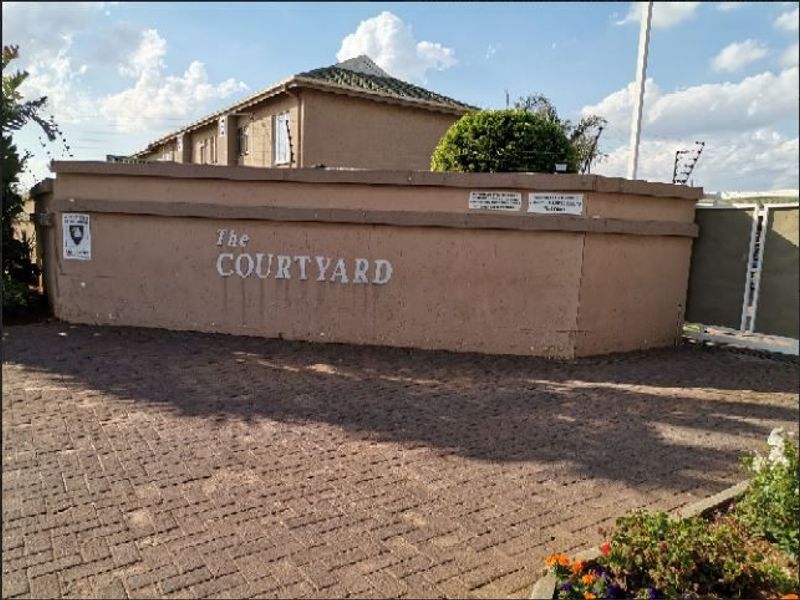 Front entrance view of The Courtyard apartment complex featuring a paved driveway, perimeter wall with electric fencing, and security gate access.