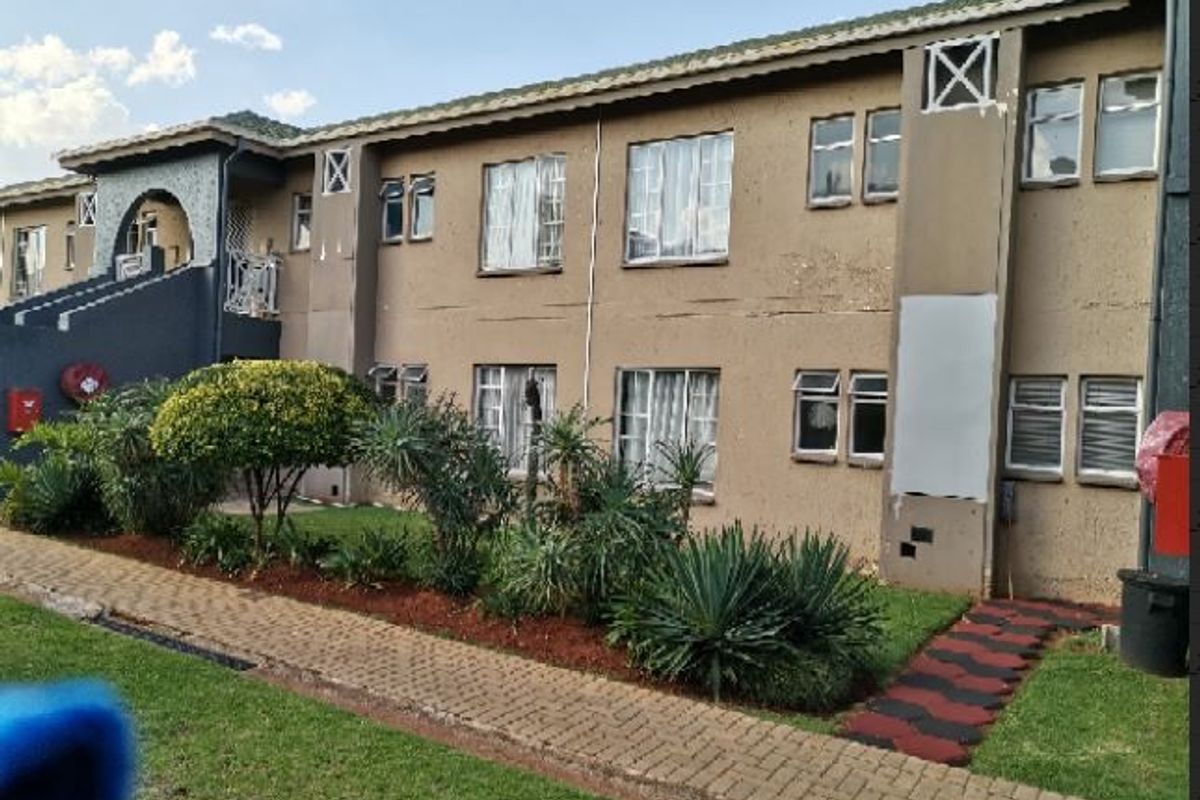 Rear exterior view of a multi-story apartment building featuring a paved walkway, landscaped garden, and visible wear to the exterior wall plaster and paint.