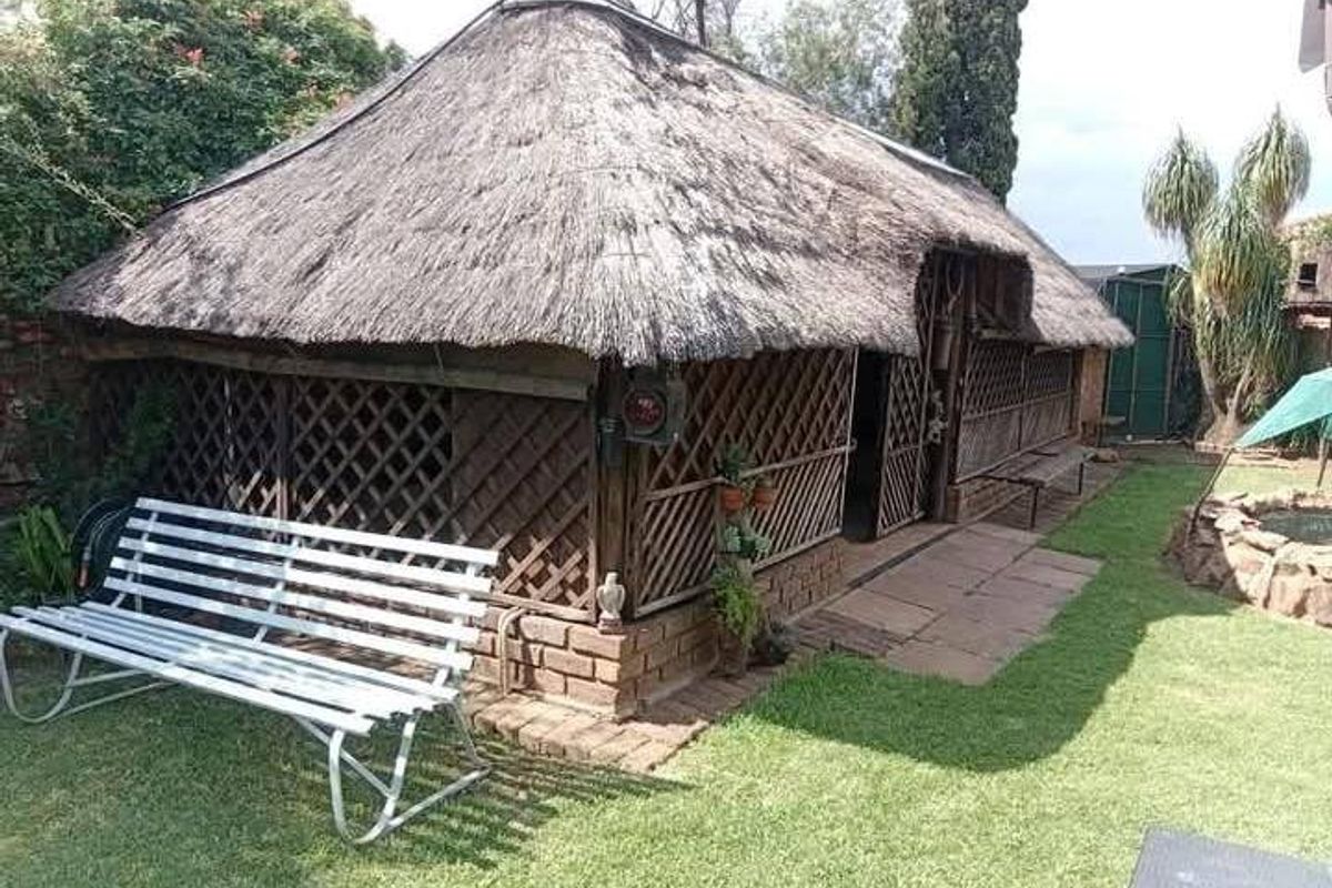 A rustic outdoor lapa structure featuring a traditional thatch roof and timber lattice walls, situated in a garden setting.