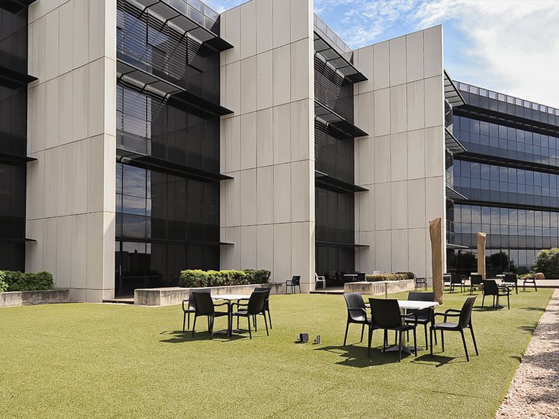 A modern commercial outdoor terrace featuring artificial turf, concrete landscaping elements, and a contemporary glass facade with integrated sun shading.