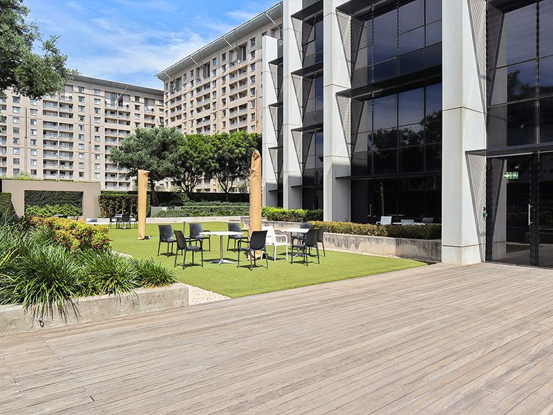 A modern commercial outdoor patio area featuring timber decking, artificial turf, and landscaped concrete planters adjacent to a glass-fronted office building.