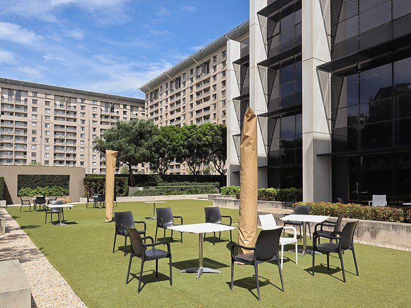 A well-maintained outdoor patio area featuring artificial turf, concrete retaining walls, and direct access to a modern commercial building with a glass facade.