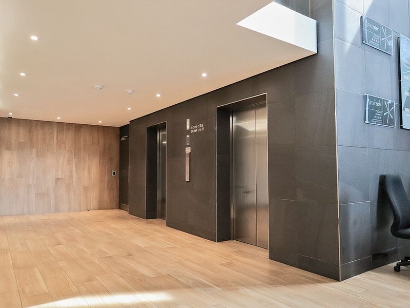 Modern commercial building lobby featuring stainless steel elevator banks, high-quality stone and wood wall finishes, and recessed ceiling lighting.