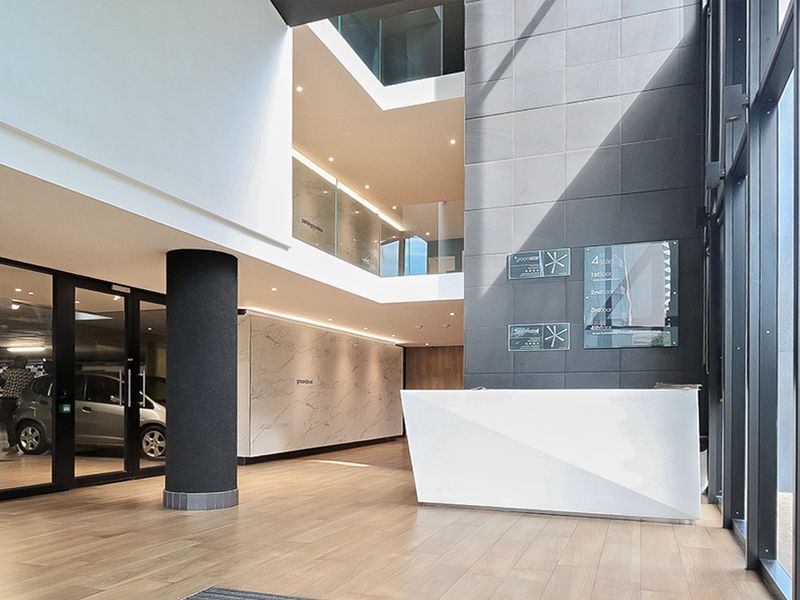 Modern commercial building lobby featuring a sleek reception desk, floor-to-ceiling glass windows, marble-look wall finishes, and a multi-level atrium design.