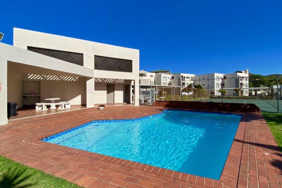 Communal swimming pool area featuring brick paving, a covered patio structure, and adjacent tennis court facilities within the apartment complex.