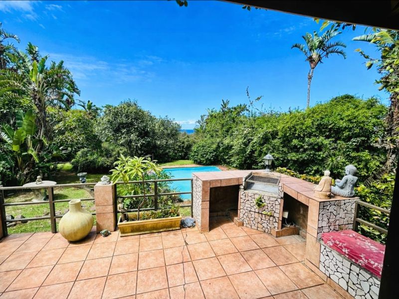 Covered patio featuring a built-in braai area, tiled flooring, and metal railings, overlooking a private swimming pool and lush garden.