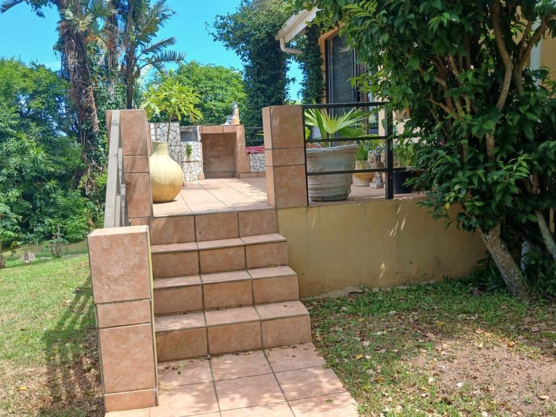 An elevated outdoor patio area featuring tiled steps, a built-in masonry braai or barbecue station, and metal railings, overlooking a garden.