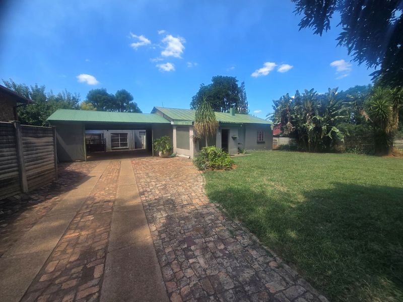 Front view of a single-story house featuring a green corrugated metal roof, a brick-paved driveway, and an attached carport set on a spacious lawn.
