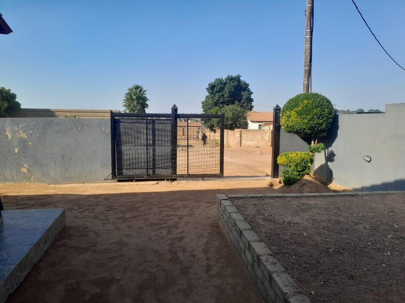 Front entrance area featuring a metal sliding gate, perimeter walling, and a concrete block retaining wall with an unpaved sandy yard.