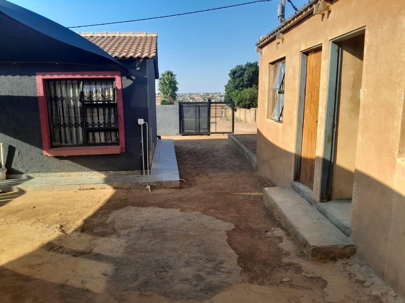 Rear exterior view showing the space between two structures with unpaved ground, security features, and basic building finishes.