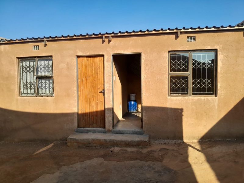 Exterior view of a single-story dwelling featuring plastered walls, metal security bars on windows, and a corrugated metal roof.