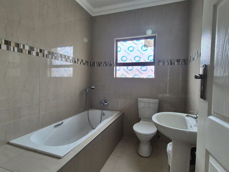 A clean, modern bathroom featuring a built-in bathtub, pedestal basin, and toilet with neutral wall and floor tiling.
