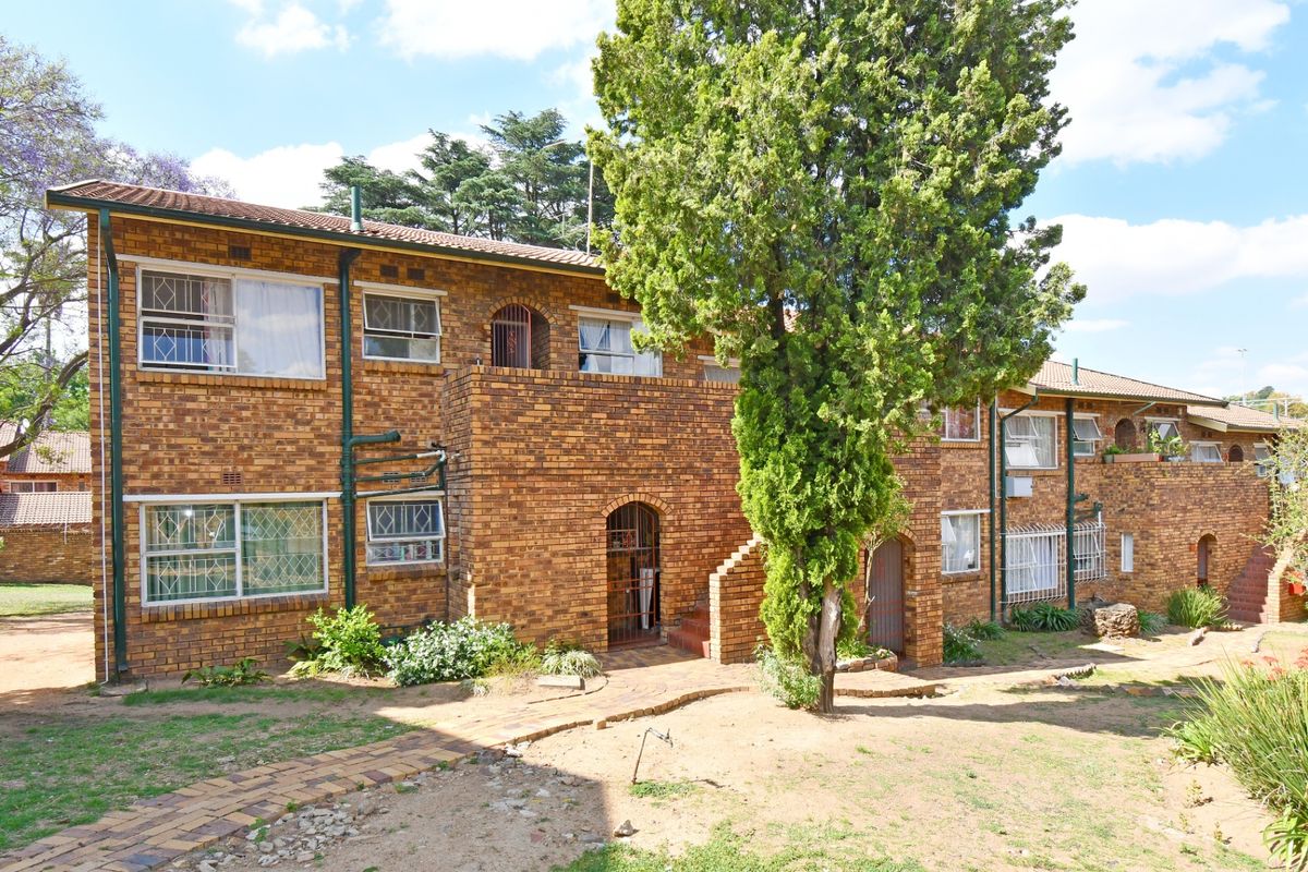 Rear view of a multi-story face-brick townhouse complex featuring established trees, paved walkways, and security-fitted windows.