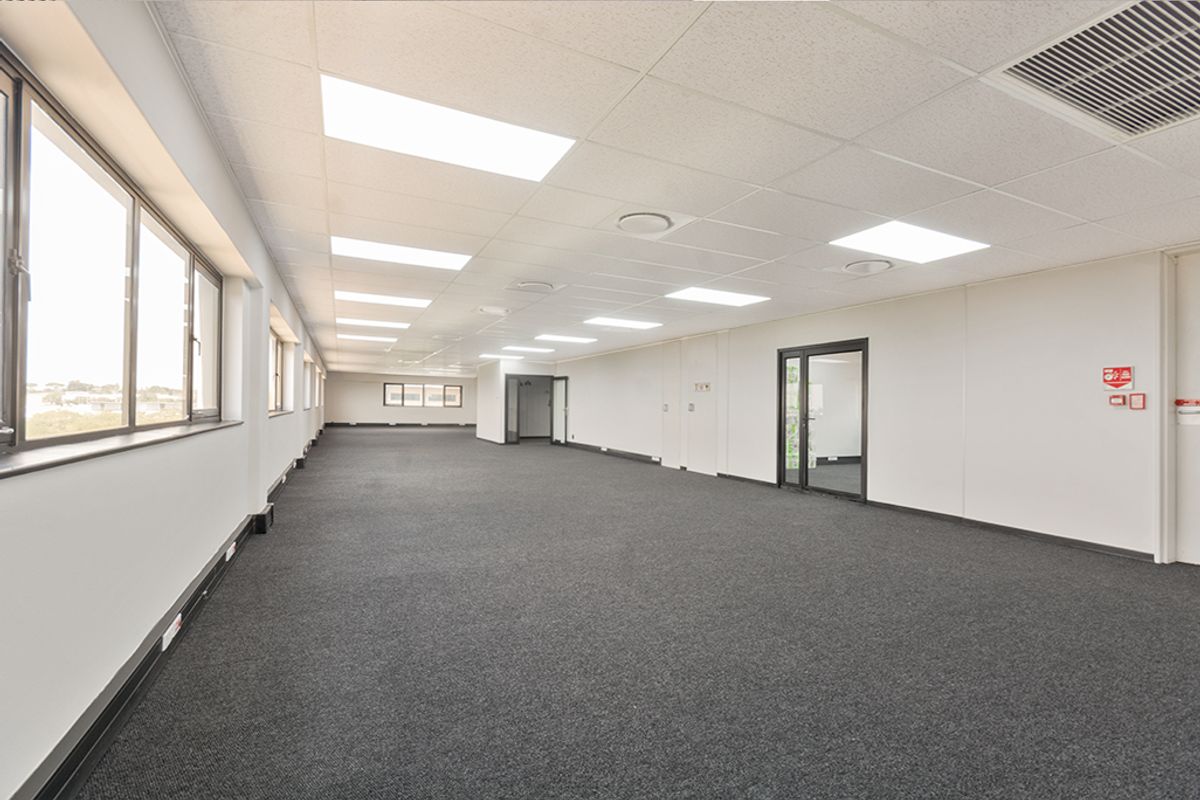 A spacious, open-plan commercial office area featuring neutral finishes, carpeted flooring, recessed lighting, and large windows providing ample natural light.
