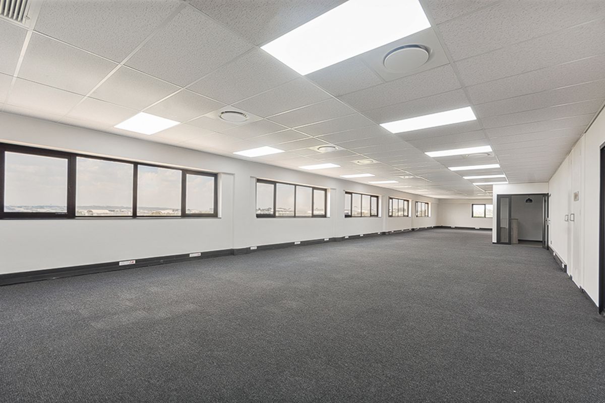 A spacious, open-plan commercial office area featuring neutral finishes, large windows providing natural light, suspended ceilings, and carpeted flooring.