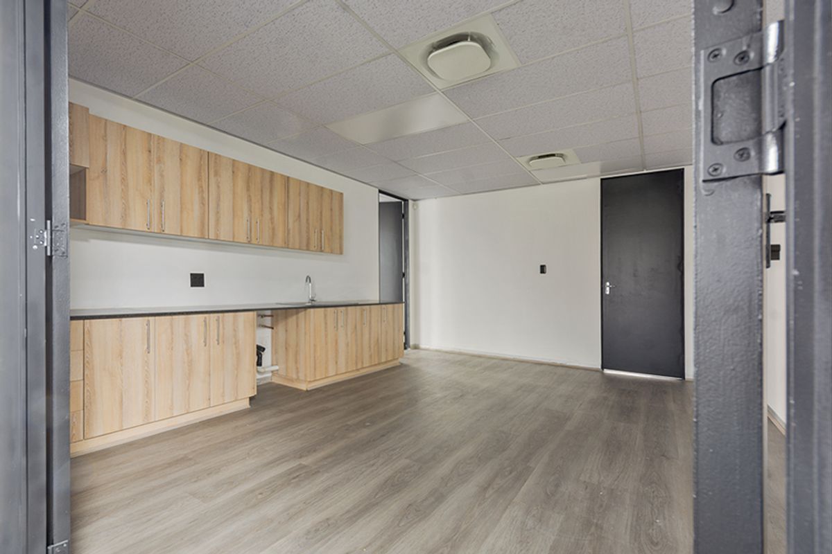 A clean, empty commercial kitchenette featuring light wood-finish cabinetry, dark countertops, a sink, and modern laminate flooring under a drop ceiling.