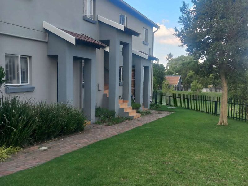 A view of the rear exterior of the townhouse, featuring a paved walkway, landscaped lawn, and metal perimeter fencing.