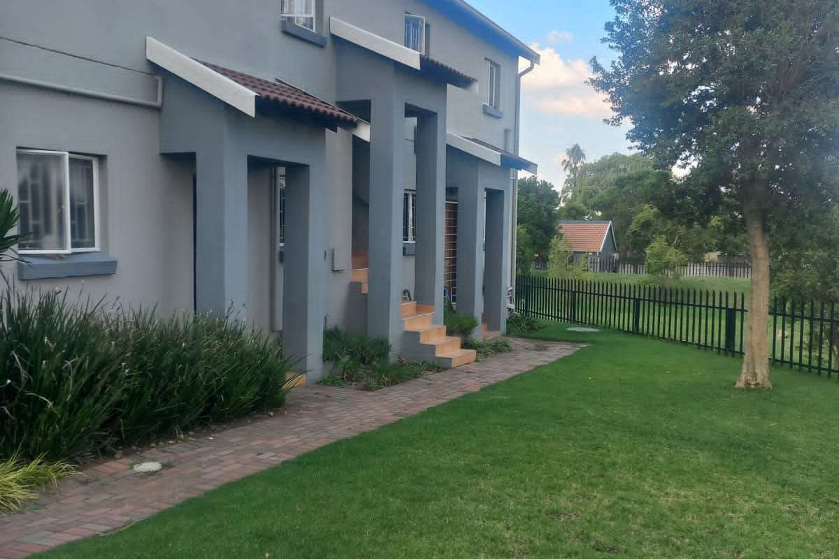 A view of the rear exterior of the townhouse, featuring a paved walkway, landscaped lawn, and metal perimeter fencing.