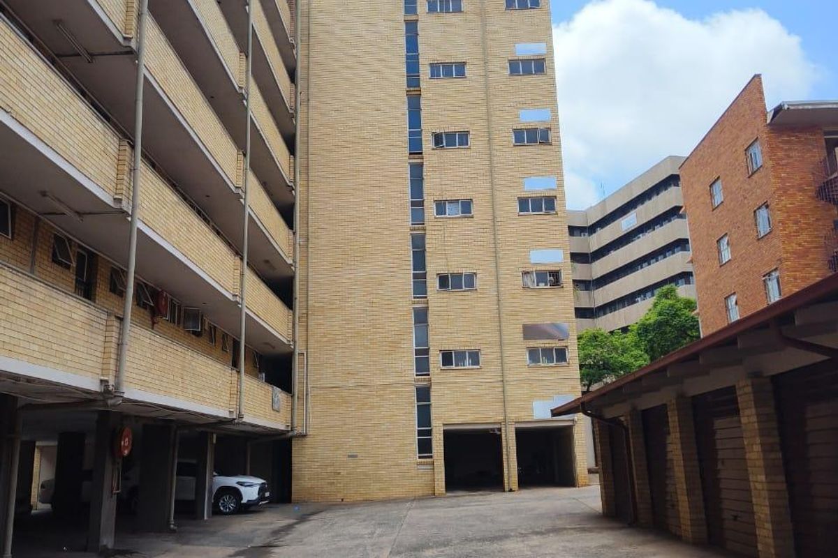 Exterior view of a multi-story apartment building featuring brick facade, open balconies, and a paved ground-level parking area with covered bays.