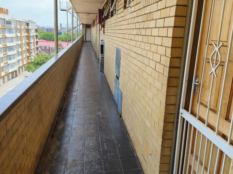 An exterior apartment balcony featuring brick walls, tiled flooring, and a metal security gate, providing access to the unit entrance.