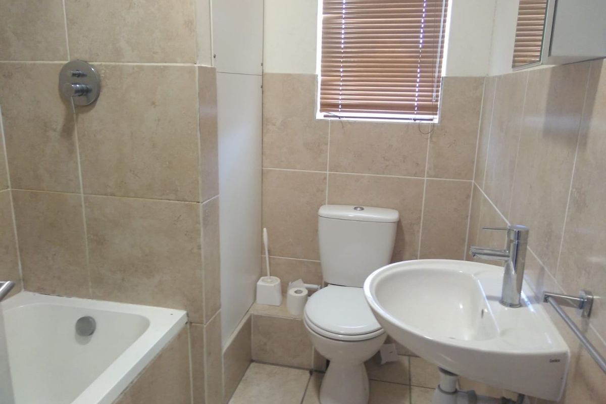 A functional bathroom featuring neutral wall and floor tiling, a standard bathtub with shower mixer, a pedestal basin, and a toilet.