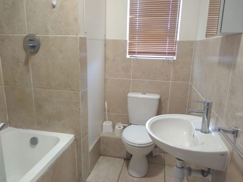 A functional bathroom featuring neutral wall and floor tiling, a standard bathtub with shower mixer, a pedestal basin, and a toilet.