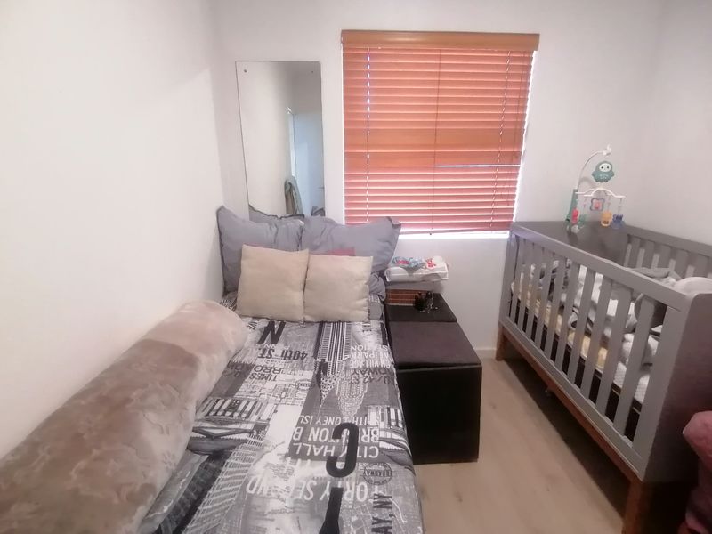 A compact bedroom featuring light-toned laminate flooring, a wall-mounted mirror, and a window fitted with wooden venetian blinds.