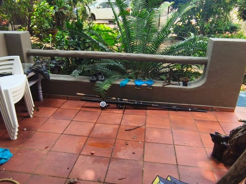 A covered patio area featuring terracotta floor tiles and a low masonry wall, overlooking a lush garden space.