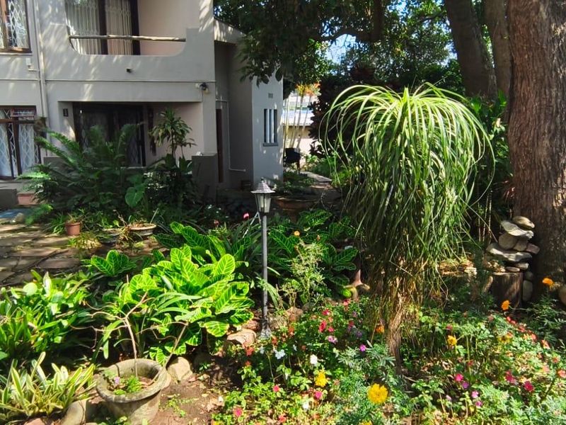 A view of the rear garden area featuring established landscaping, mature trees, and a paved pathway leading to the apartment building exterior.