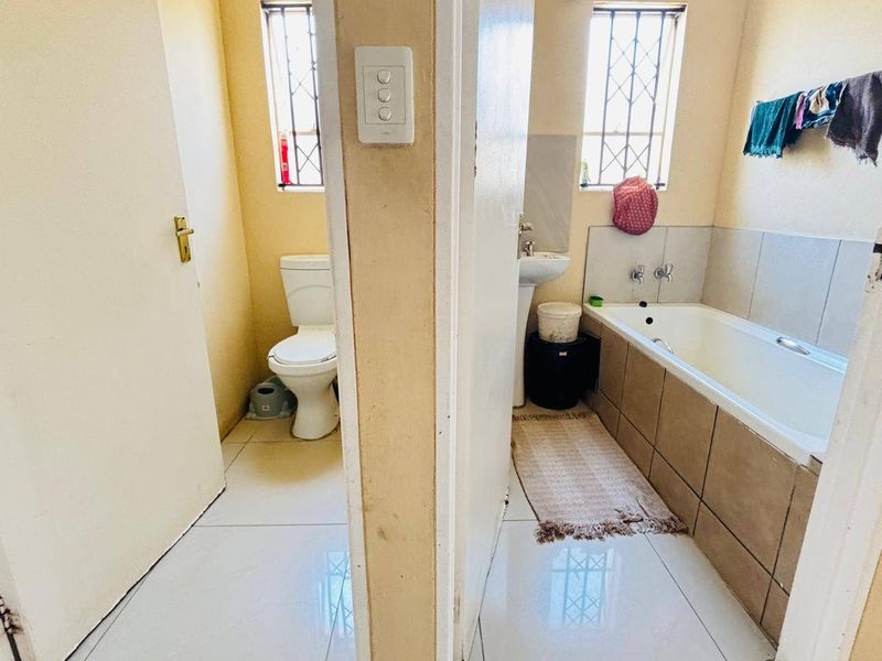 A functional bathroom featuring a separate toilet area, pedestal basin, and a tiled bathtub with security bars on the windows.