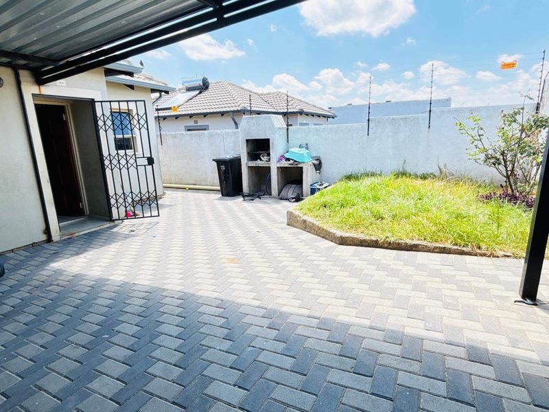 A paved outdoor patio area featuring a built-in braai, covered roofing, and a small garden section enclosed by a perimeter wall with electric fencing.