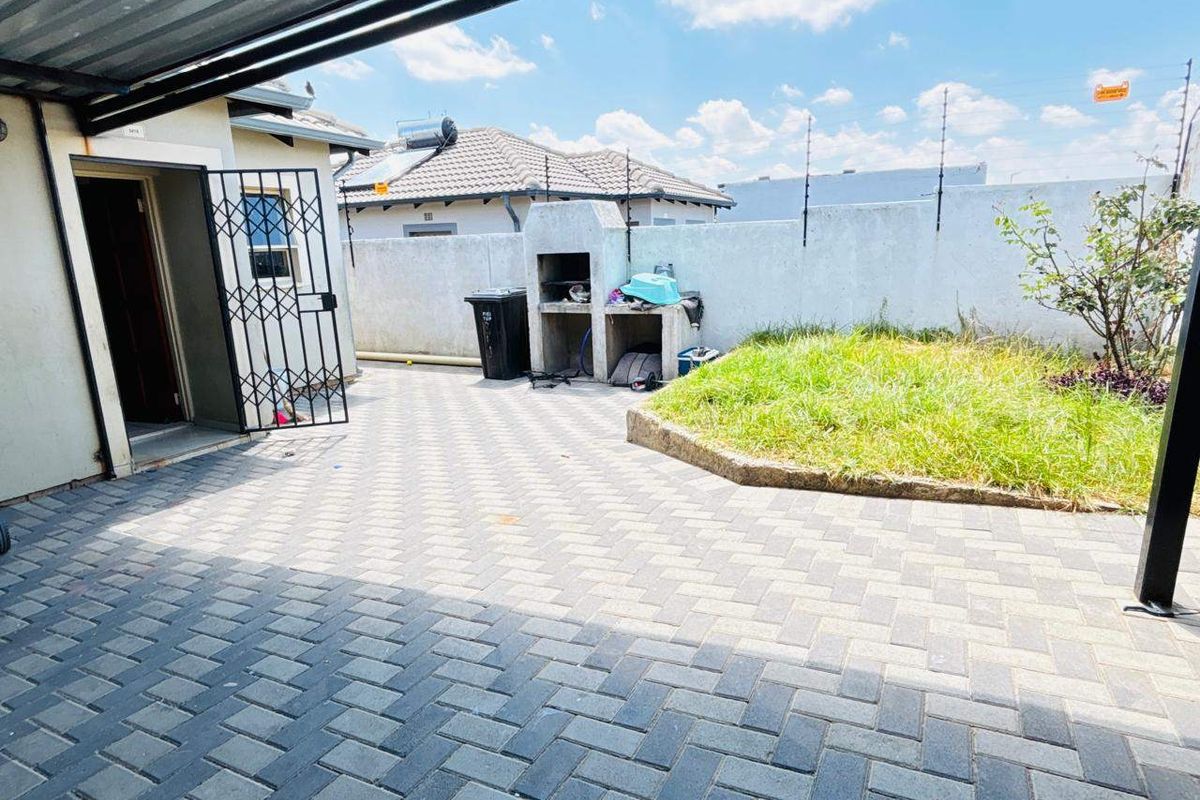 A paved outdoor patio area featuring a built-in braai, covered roofing, and a small garden section enclosed by a perimeter wall with electric fencing.