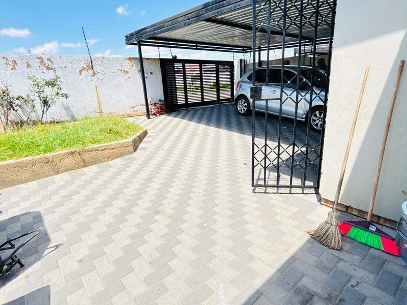 Front exterior area featuring a paved driveway, covered carport, and secure metal gate access with a perimeter wall.