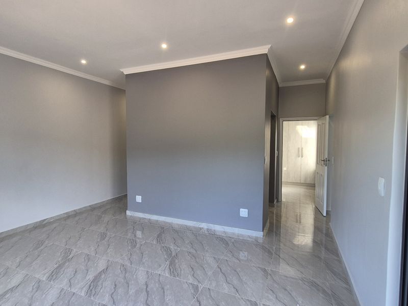 A bright, empty living area featuring modern grey-toned porcelain floor tiles, recessed downlighting, and coved cornicing, leading to a hallway with built-in storage.