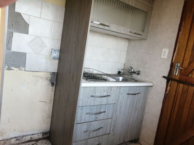 A small kitchen area featuring built-in cabinetry and a sink, currently requiring significant wall repairs and tiling to restore the finish.