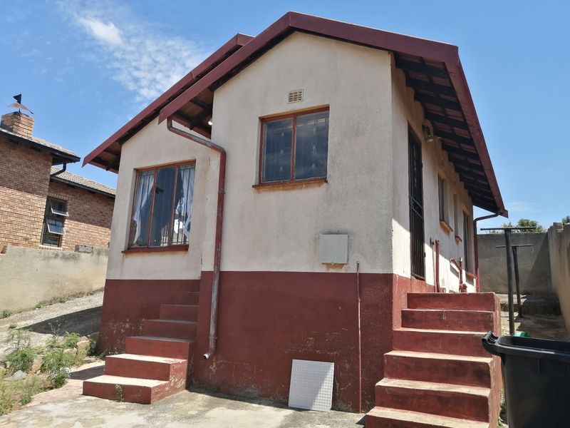 Rear exterior view of a detached house featuring concrete stairs, plastered walls, and a pitched roof with visible signs of wear.