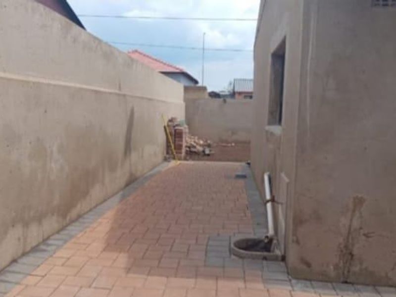 A narrow side passage featuring paved flooring, a high boundary wall, and exterior drainage infrastructure along the side of the house.