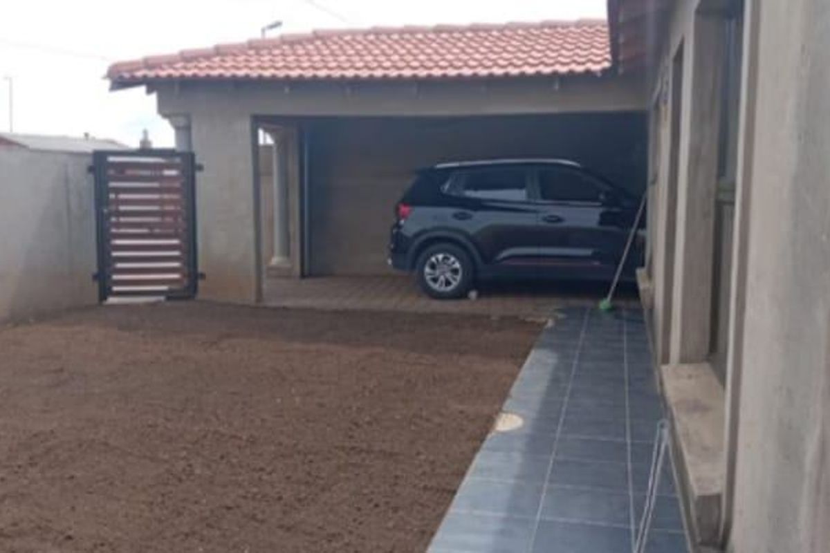 Rear exterior view showing a covered patio area, paved driveway, and boundary wall with an unfinished garden space.