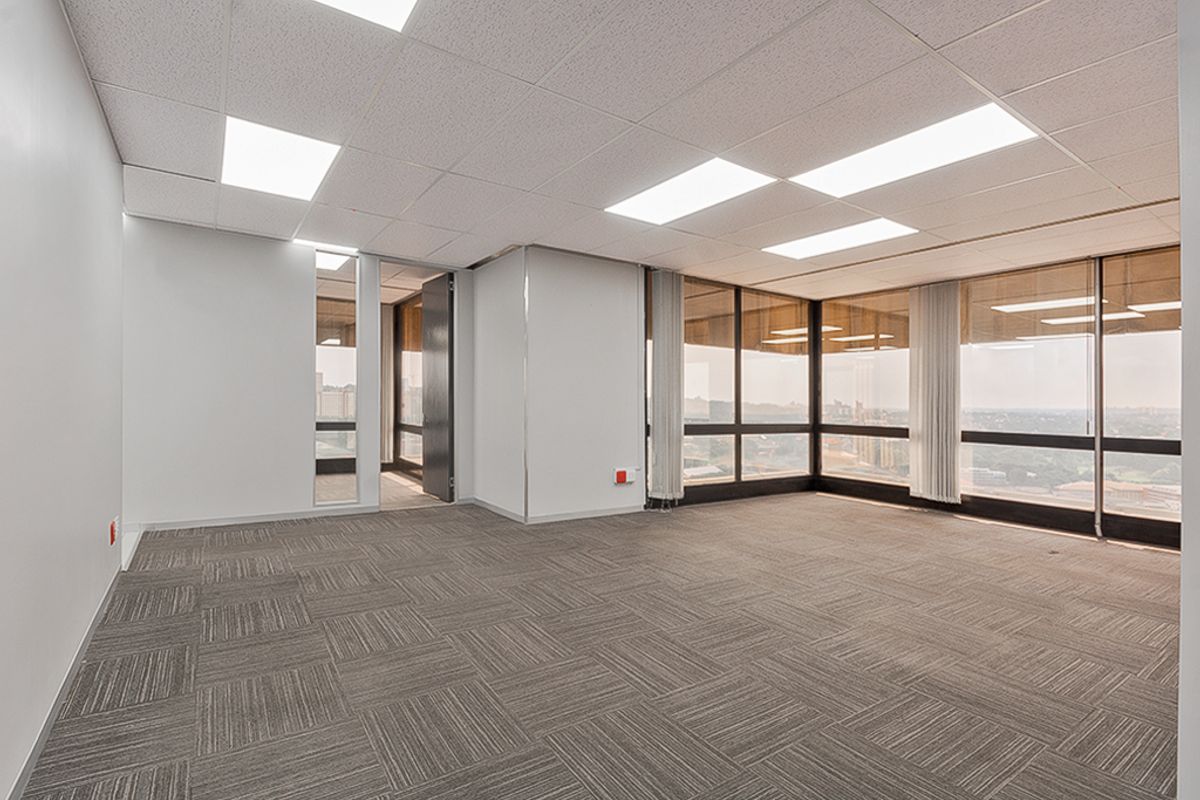 A bright, vacant commercial office space featuring carpet tiles, suspended ceilings with recessed lighting, and expansive floor-to-ceiling windows offering panoramic views.