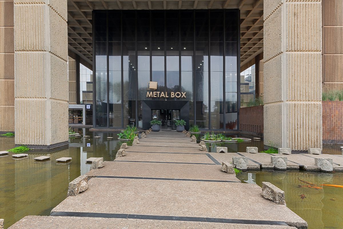 The main entrance of the Metal Box commercial building, featuring a prominent glass facade, concrete structural pillars, and a decorative water feature with a walkway.
