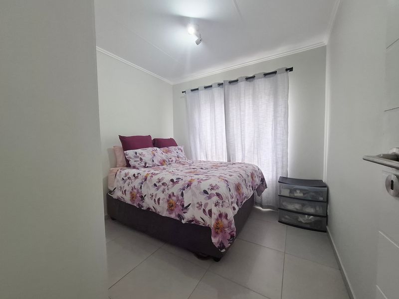 A compact bedroom featuring neutral tiled flooring, white walls, and a window dressed with curtains.
