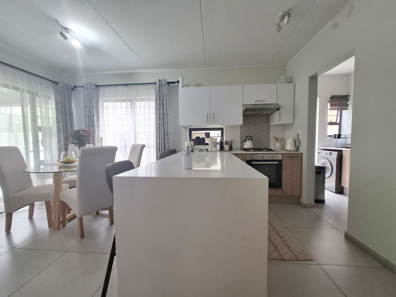 Modern open-plan kitchen and dining area featuring a central island, sleek cabinetry, built-in appliances, and neutral tiled flooring with access to a separate laundry area.