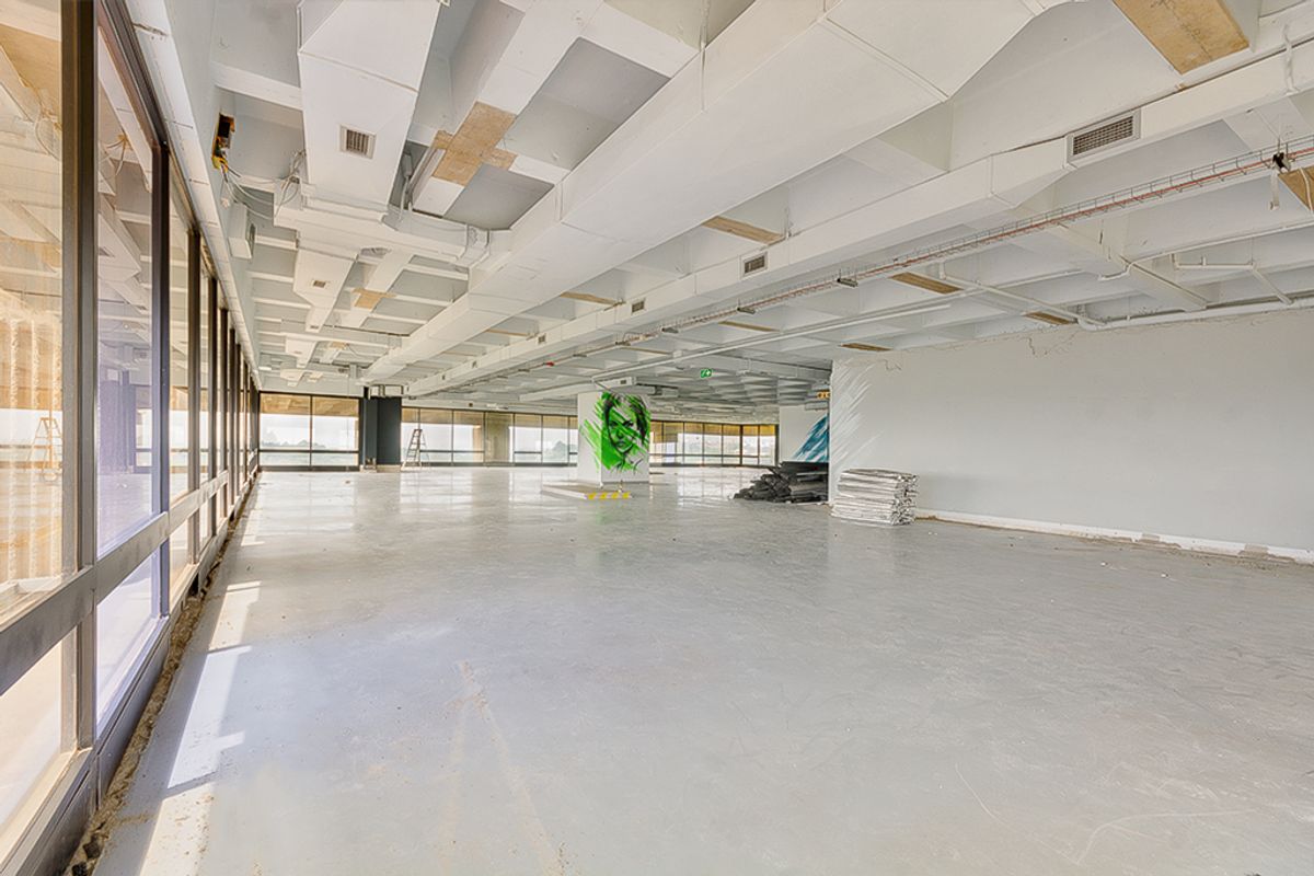 A large, open-plan commercial office space featuring exposed ceiling ductwork, polished concrete floors, and extensive floor-to-ceiling windows providing ample natural light.