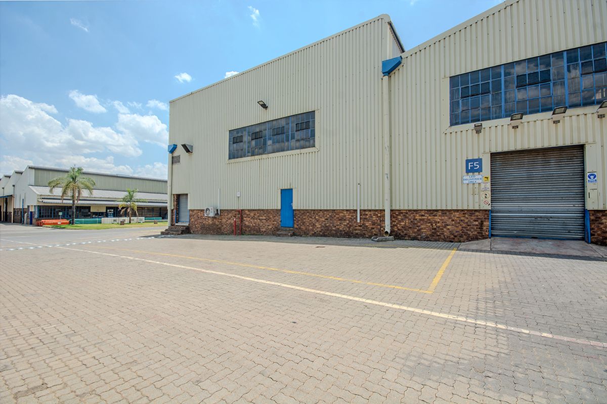 Industrial warehouse exterior featuring corrugated metal cladding, a brick base, paved yard area, and a large roller shutter door for logistics access.