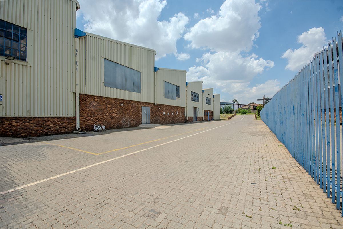 Rear exterior view of an industrial warehouse facility featuring corrugated metal cladding, face brick lower walls, and a paved driveway with secure steel palisade fencing.