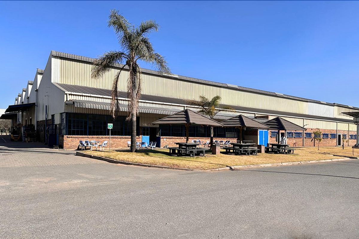 Exterior view of a large industrial warehouse facility featuring corrugated metal cladding, brick base, and paved outdoor area with fixed shade structures.