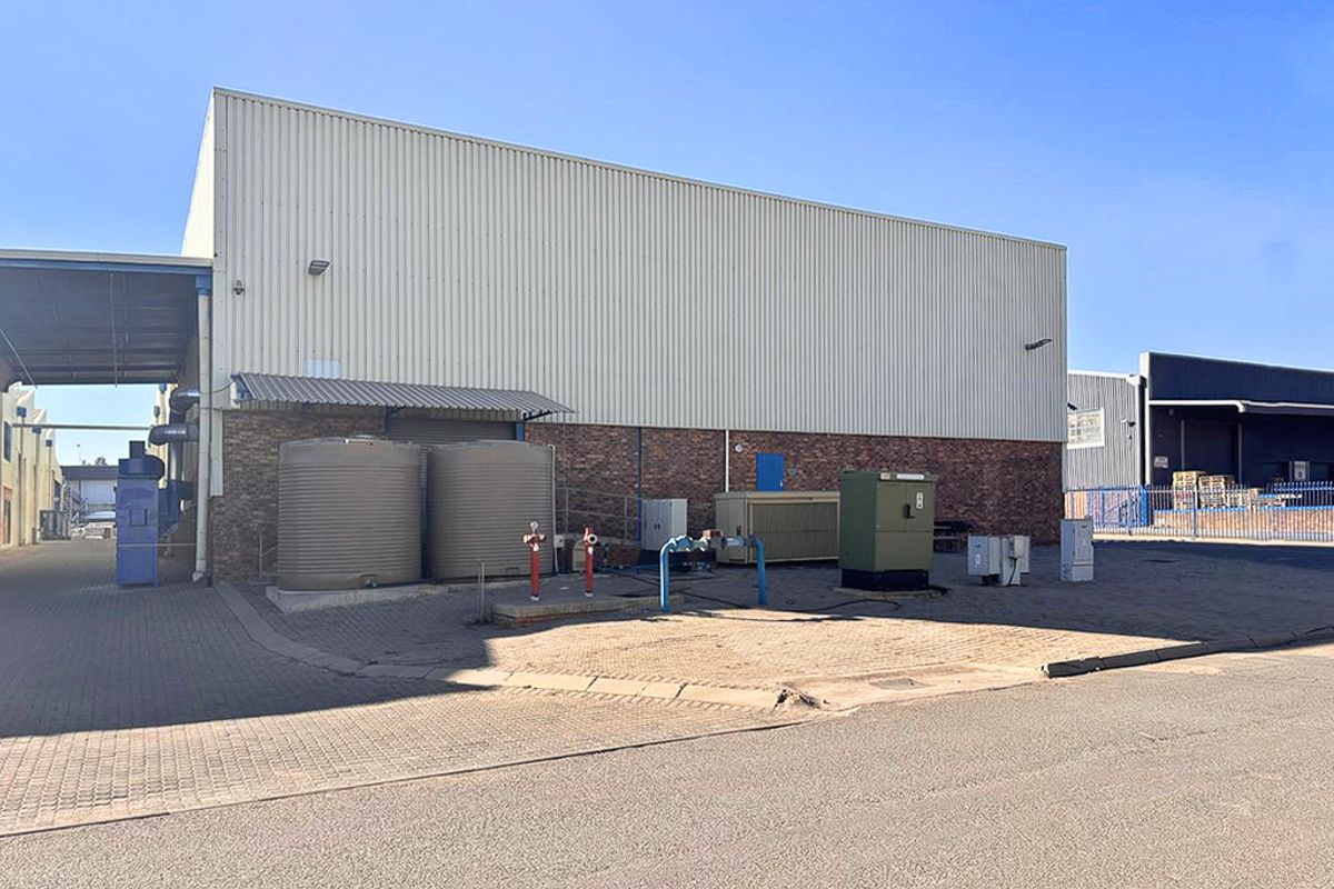 Rear exterior view of an industrial warehouse featuring corrugated metal cladding, brick base, water storage tanks, and external utility infrastructure on a paved yard.